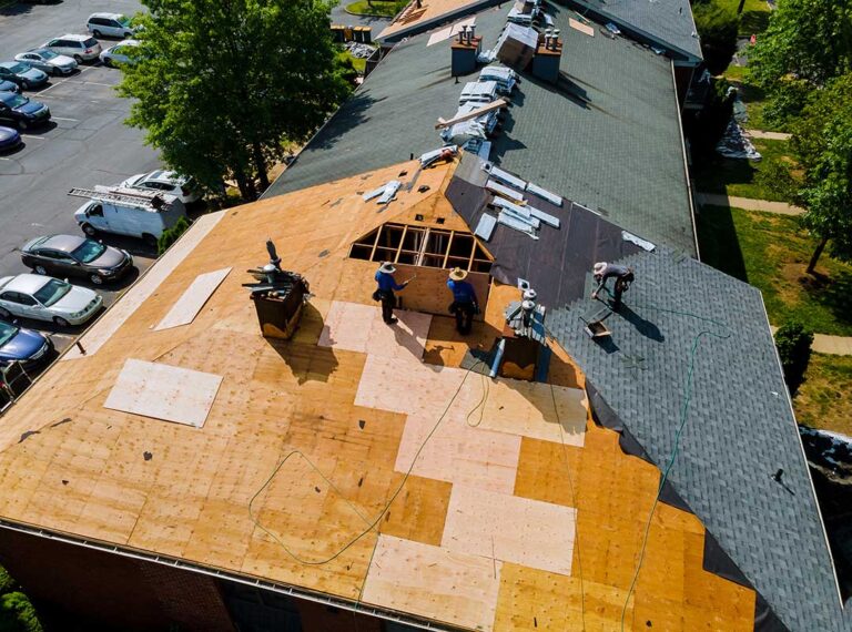 A homeowner on a ladder gently cleaning leaves from a shingle roof and clearing out gutters.